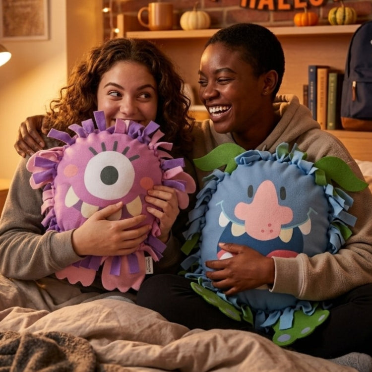 Two college students sitting together in bed holding colorful monster-shaped plush pillows in a cozy indoor setting.