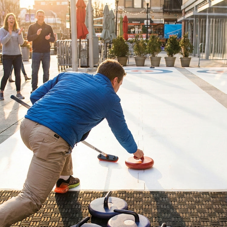Man playing curling on a synthetic ice rink at an event while two onlookers watch and cheer.