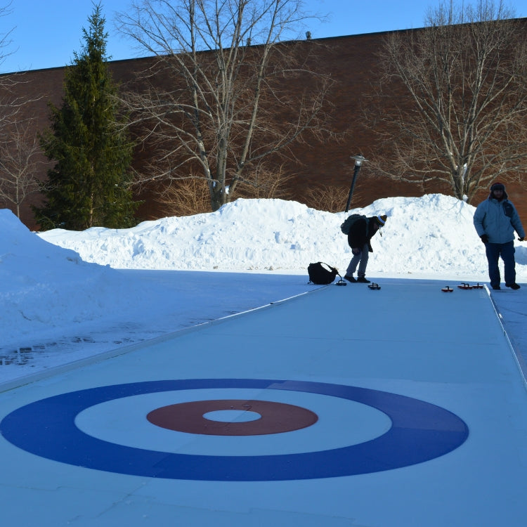 Two people on a curling rink with a large snow bank in the background.