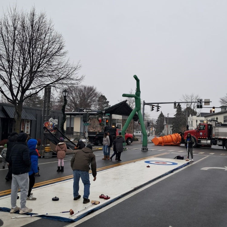 People playing curling on an outdoor rink in Upstate New York with a street and traffic lights in the background.