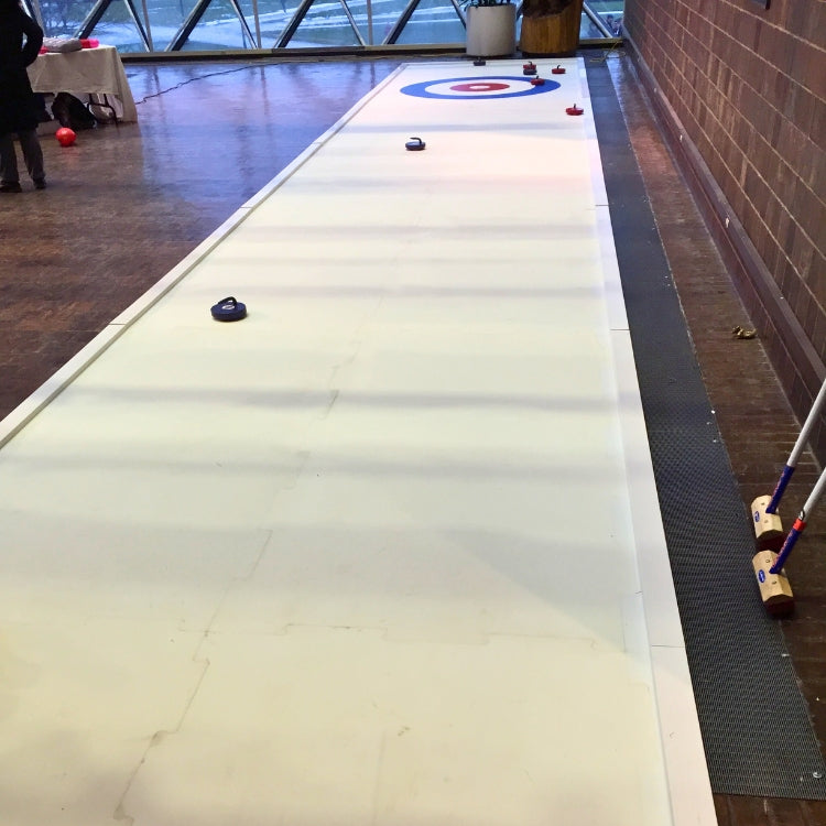 Curling rink rental setup indoors with curling brooms and stones on a polished wooden floor in Rochester, NY.