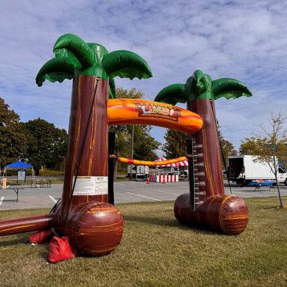 Inflatable Limbo game rental with palm tree design at an event on a grassy area with trees and a blue sky in the background.